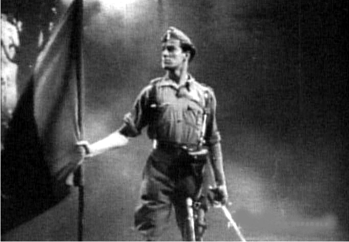 Fotograma de la película propagandística Rojo y negro (1942) en la que un soldado de uniforme porta la bandera falangista. Imagen: CEPICSA.