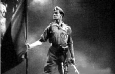 Fotograma de la película propagandística Rojo y negro (1942) en la que un soldado de uniforme porta la bandera falangista. Imagen: CEPICSA.